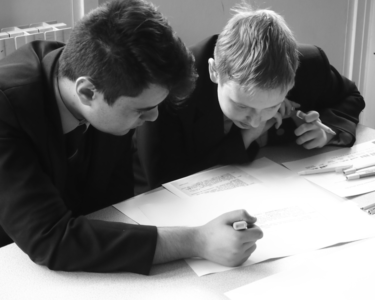 Black‑and‑white photograph of two students in school uniforms sitting at a desk, concentrating on written work. One student holds a pen and points to text on a sheet of paper, while the other leans in closely to read. Several papers and pens lie scattered on the table, suggesting collaborative study or discussion.