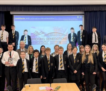 Group photo of secondary school students and staff standing on a stage and in front of it, dressed in black blazers with striped ties. Behind them, a projected slide reads “Lesson Objective: Reflect on the importance of Holocaust Memorial Day” and “Bridging Generations.” The setting is a school hall with a blue curtain backdrop.