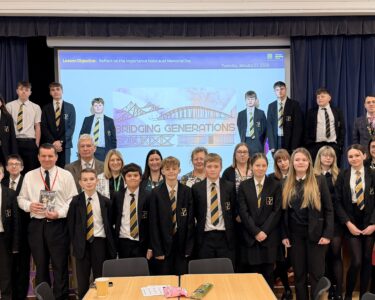 Group photo of secondary school students and staff standing on a stage and in front of it, dressed in black blazers with striped ties. Behind them, a projected slide reads “Lesson Objective: Reflect on the importance of Holocaust Memorial Day” and “Bridging Generations.” The setting is a school hall with a blue curtain backdrop.