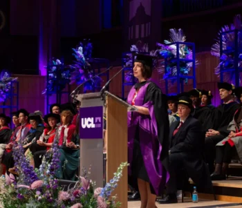 A formal graduation ceremony at UCL. A speaker in a purple academic gown and mortarboard stands at a podium delivering a speech. Behind them, rows of faculty and graduates in academic regalia are seated. The stage is decorated with flowers and lit with purple lighting.